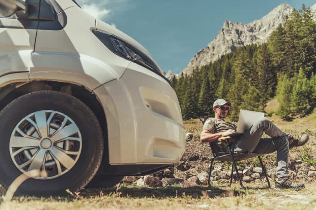 A man working as a virtual assistant on his laptop outside next to his van.